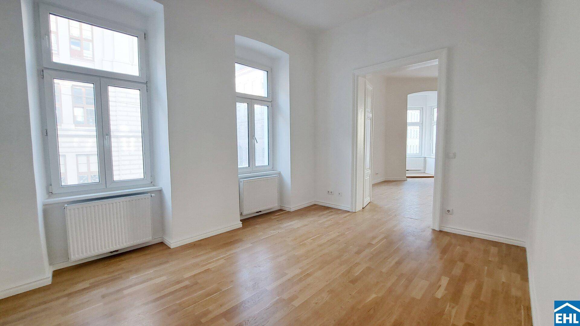 Heller Raum mit Parkettboden, hohen Fenstern und einem weiten Durchgang zu einem angrenzenden Zimmer.
