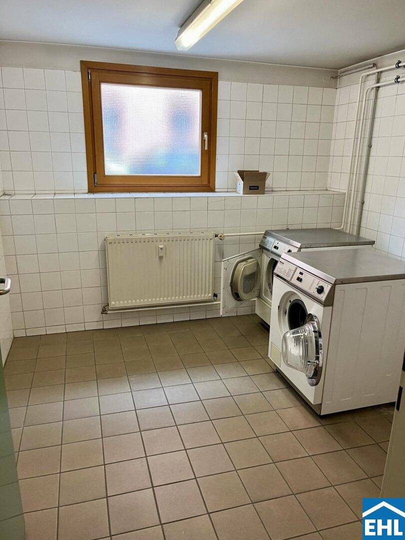 Gemeinschaftlicher Waschraum mit Waschmaschinen, Fenster und gefliesten Wänden.