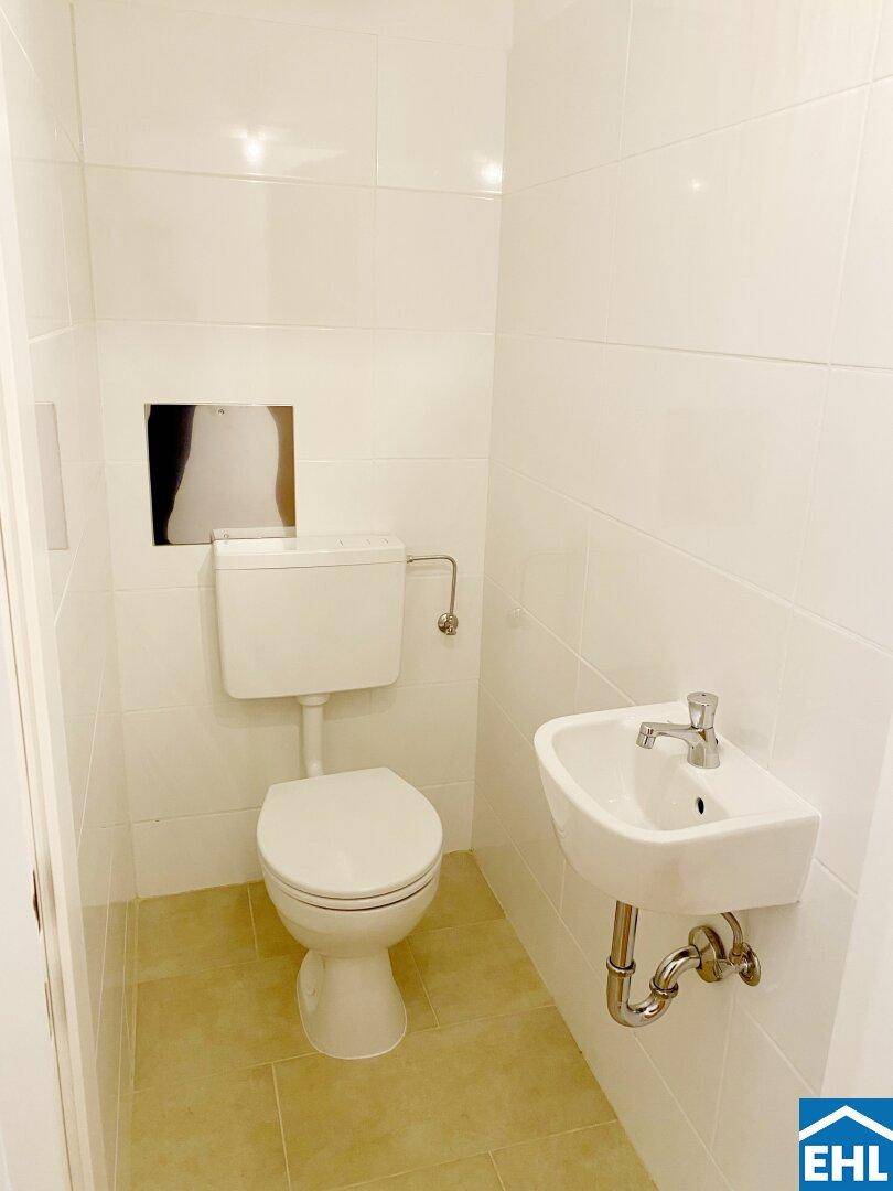 Separates WC mit Toilette, kleinem Waschbecken und weißen Wandfliesen.