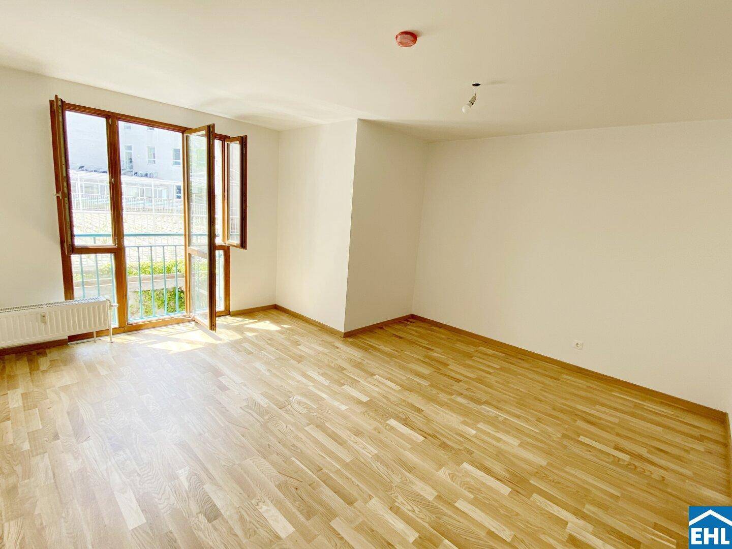 Geräumiger Raum mit hellem Parkettboden, weißen Wänden und einem französischen Balkon.
