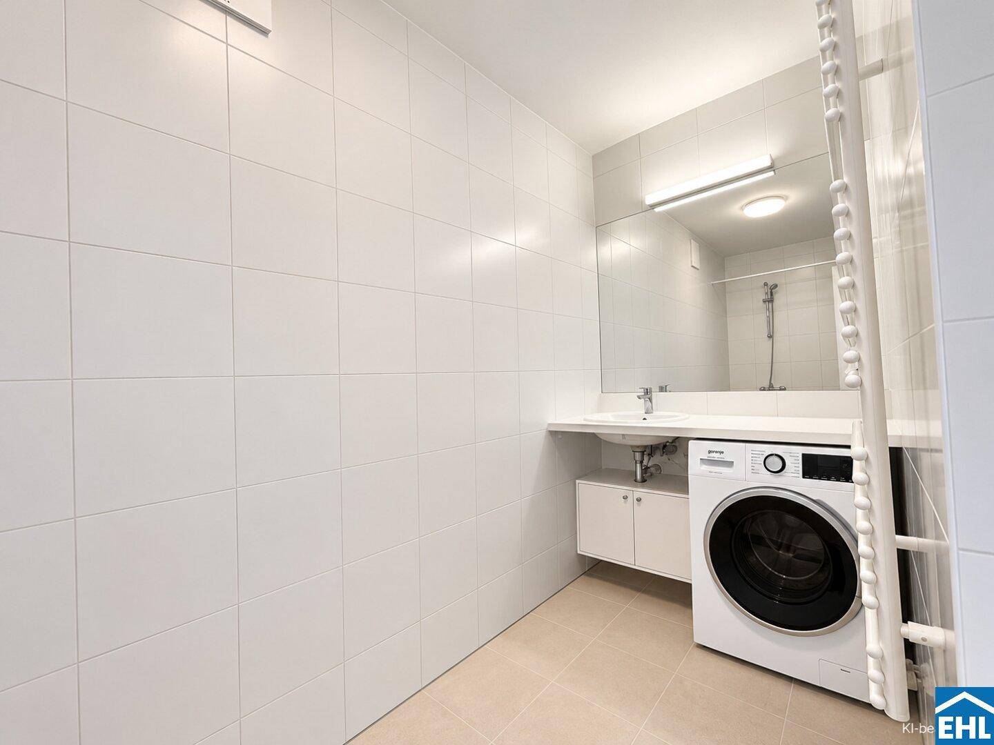 Helles Badezimmer mit weißen Fliesen, Waschbecken, Spiegel und integrierter Waschmaschine.