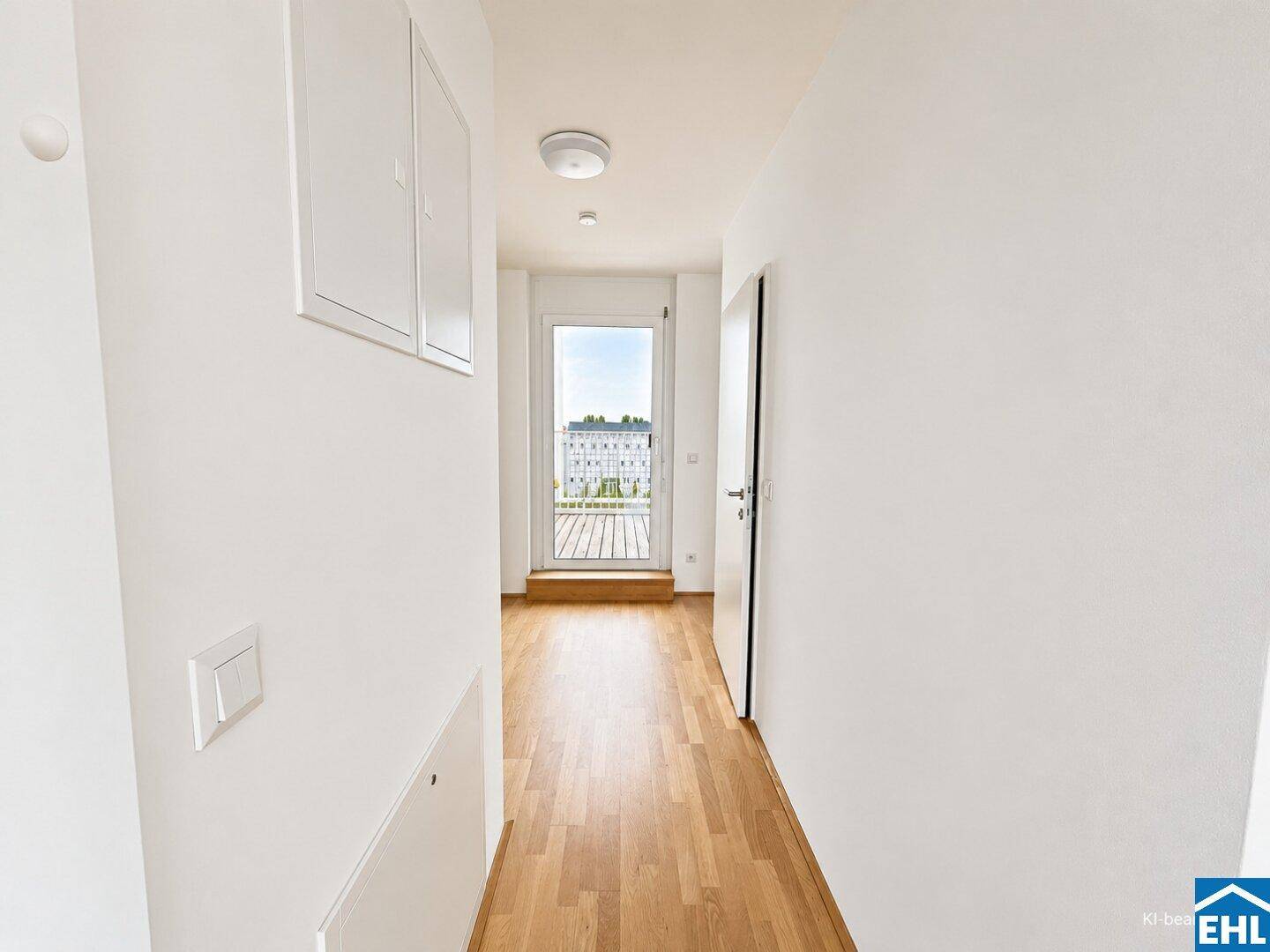 Heller Flur mit Holzboden und weißen Wänden, führt zu einem Ausgang mit Blick ins Freie.