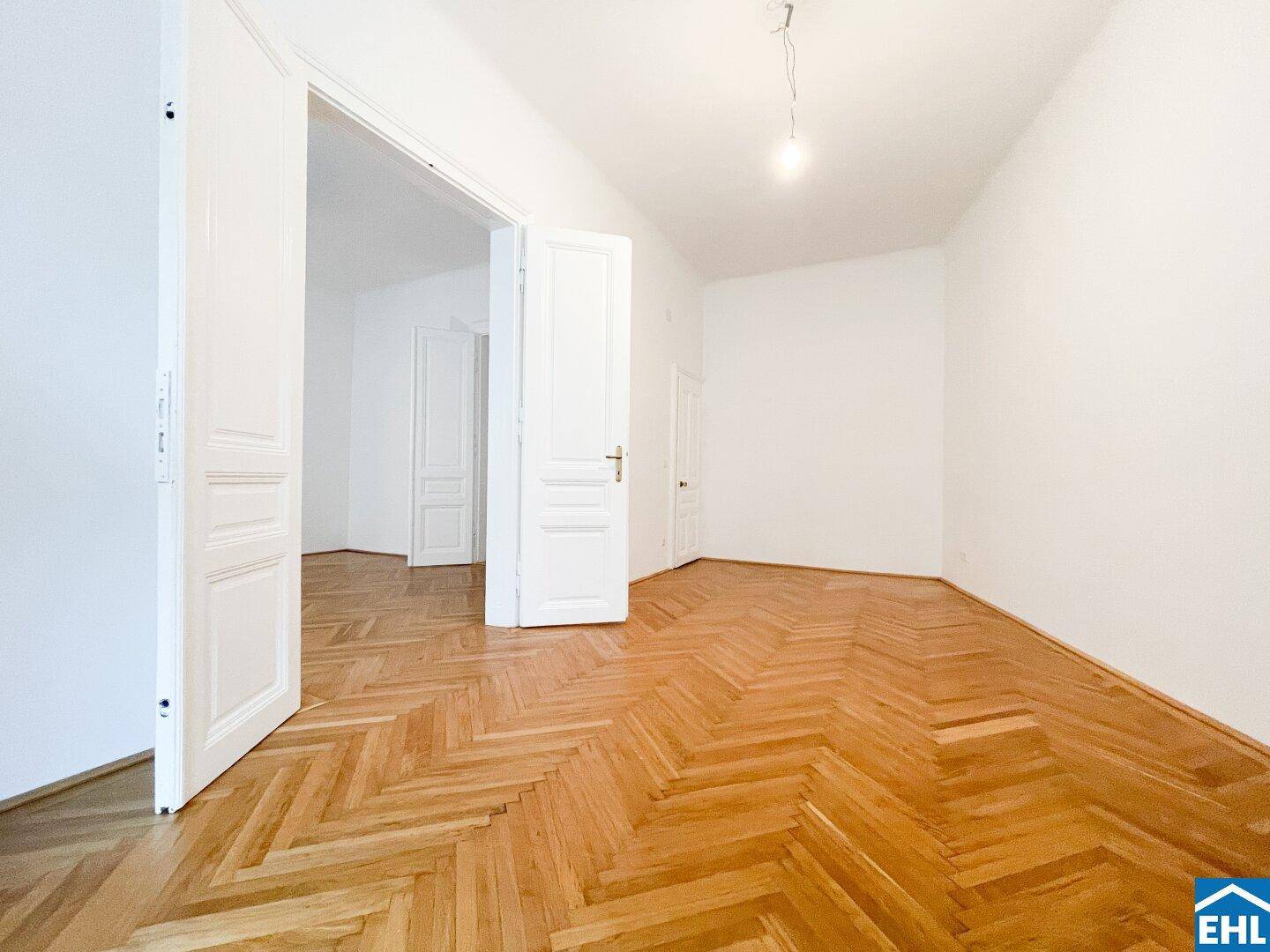 Geräumiges Zimmer mit klassischem Fischgrätparkett und hohen weißen Wänden.