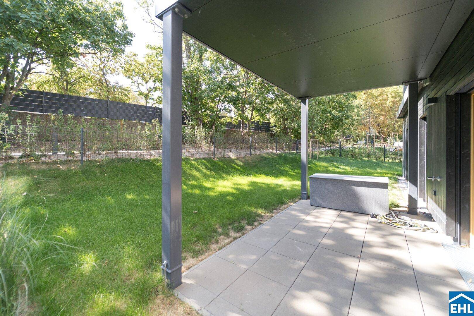 Überdachte Terrasse mit Steinplatten und angrenzendem Rasenbereich, umgeben von Bäumen.