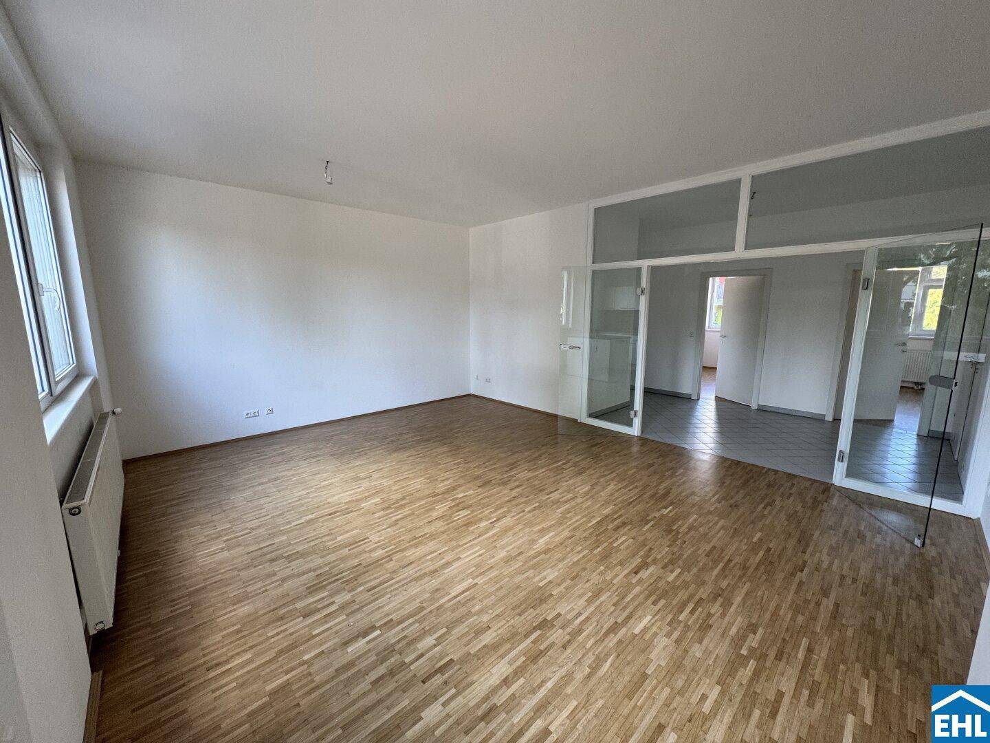 Geräumiger Wohnbereich mit Parkettboden, großen Fenstern und Zugang zu weiteren Räumen.