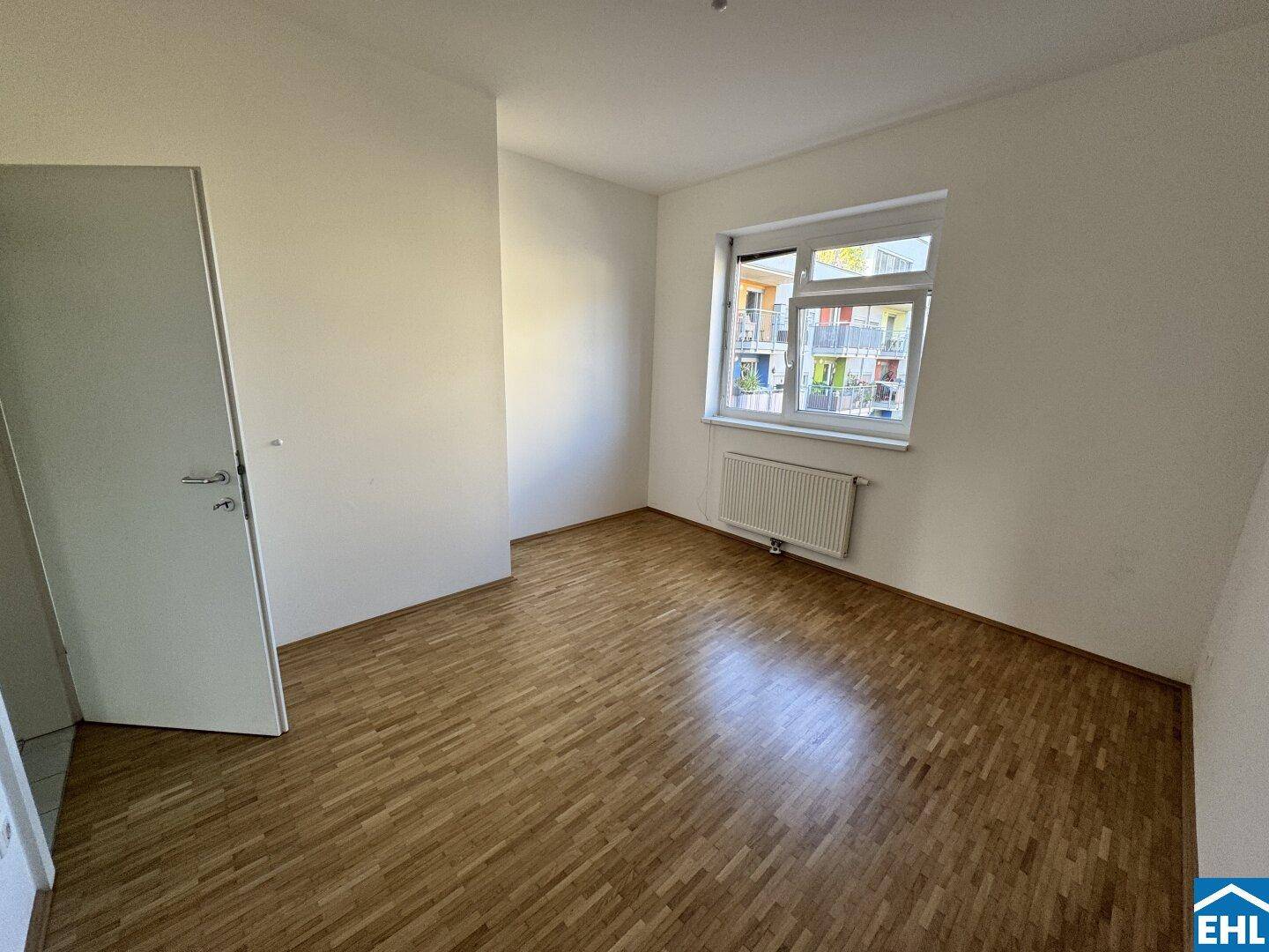 Helles Schlafzimmer mit Parkettboden, weiß gestrichenen Wänden und Blick auf die Nachbarschaft.