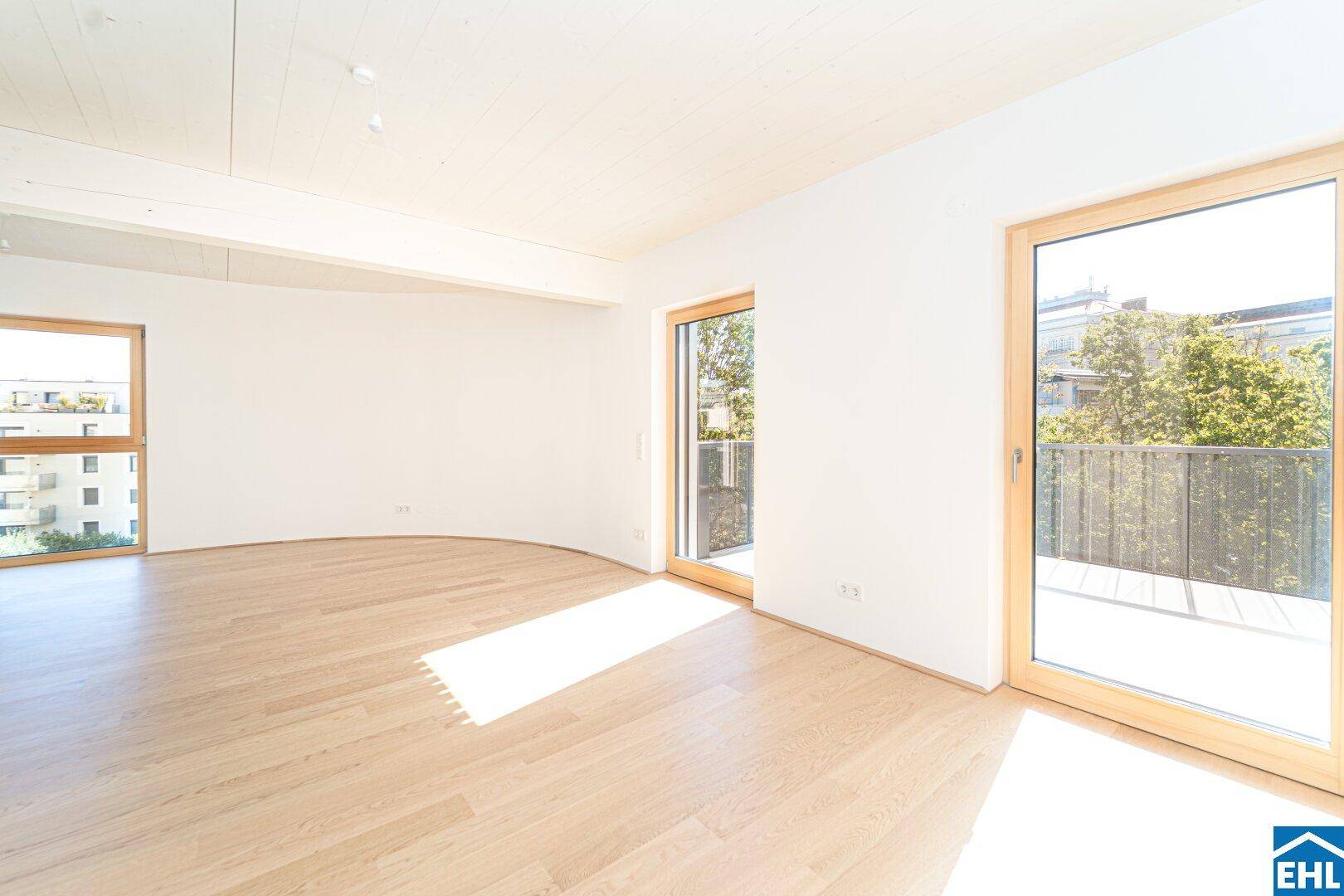Helles, leeres Zimmer mit Holzboden, weißen Wänden und Zugang zu einem Balkon, durchflutet von Sonnenlicht.