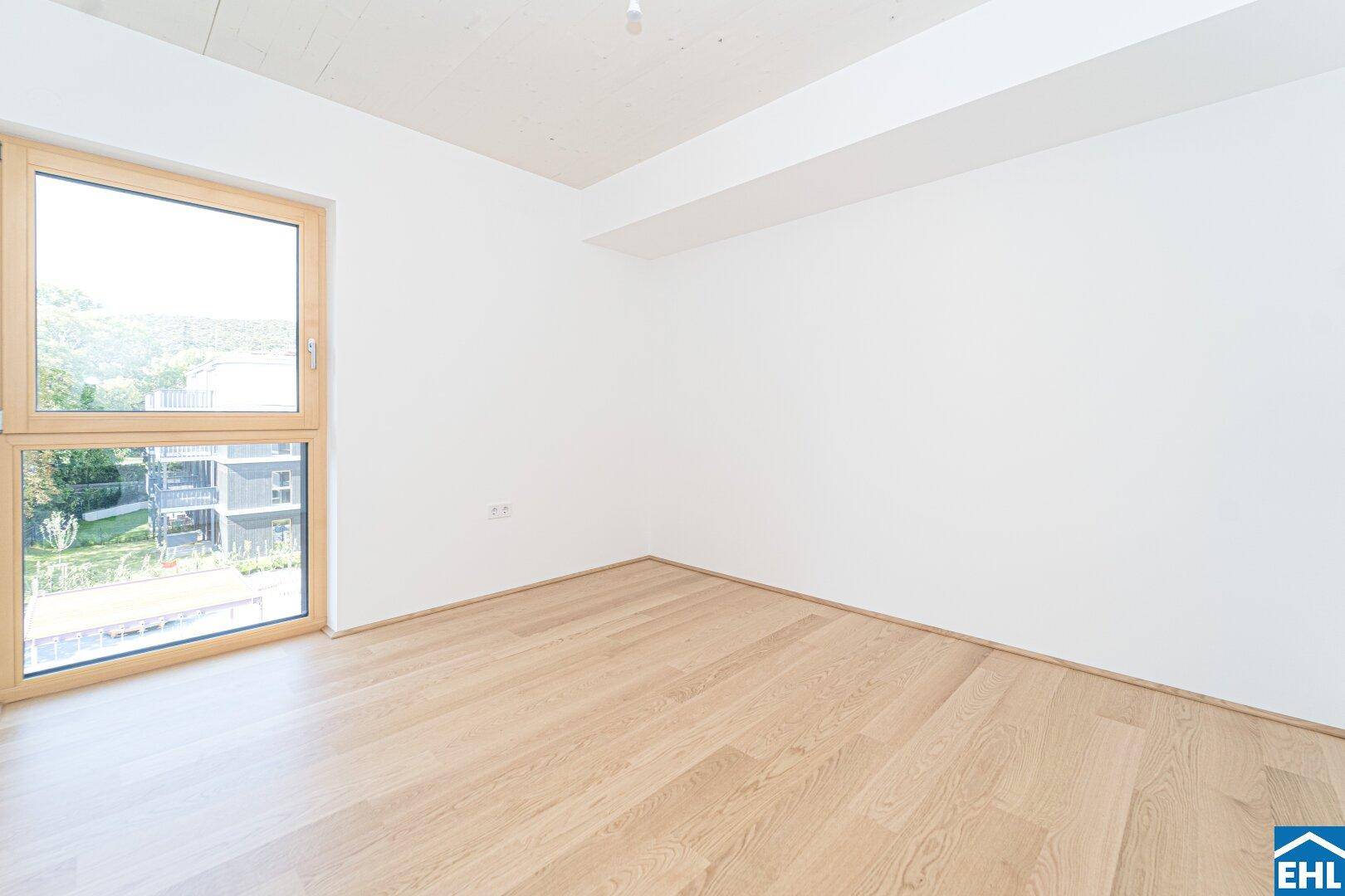 Freundliches Zimmer mit hellem Parkettboden und einem großen Fenster, das viel Licht hereinlässt.