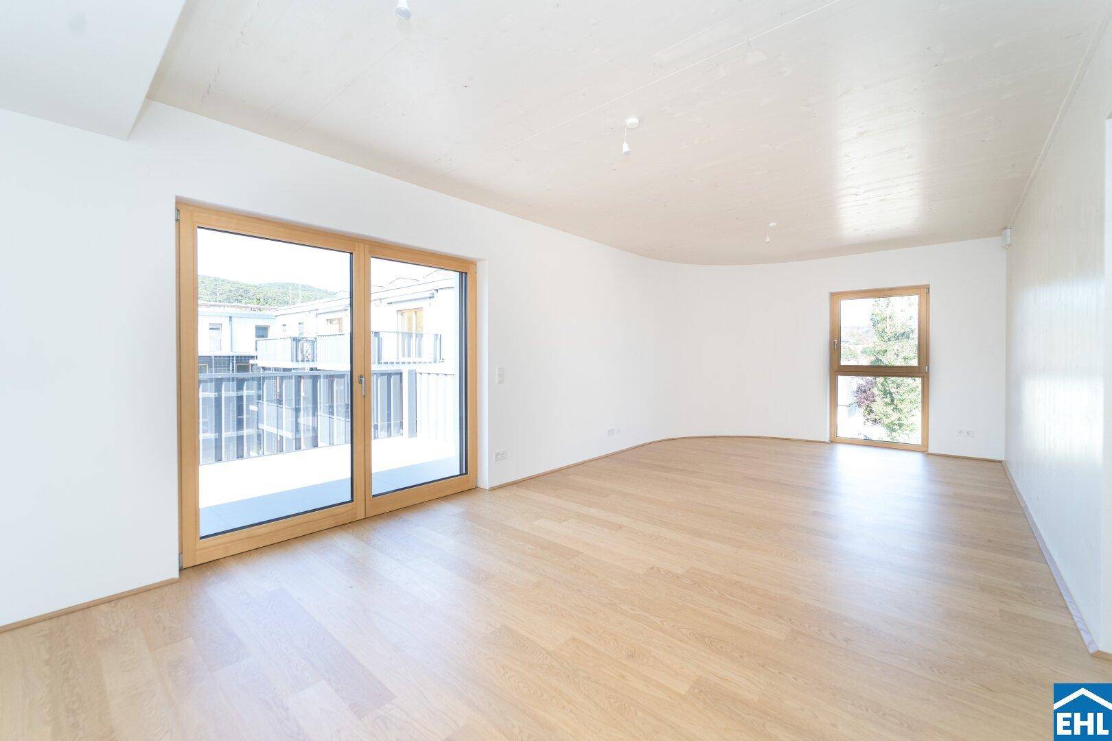 Geräumiges Wohnzimmer mit hellem Holzfußboden und großen Fenstern zum Balkon.