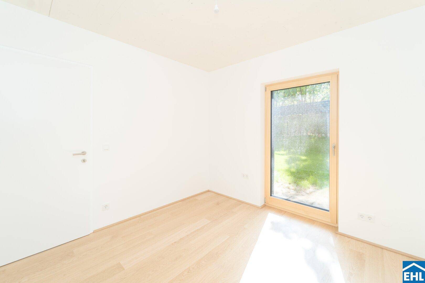 Leeres Zimmer mit Holzboden und einem Fenster, das den Blick auf den Garten freigibt.