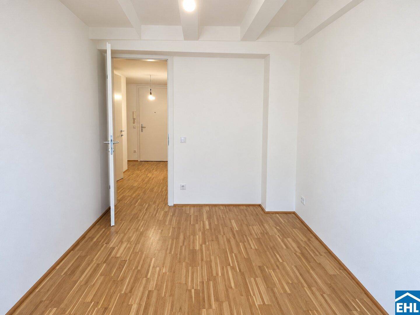 Heller Raum mit Parkettboden und offener Tür, die zu einem weiteren Bereich führt.