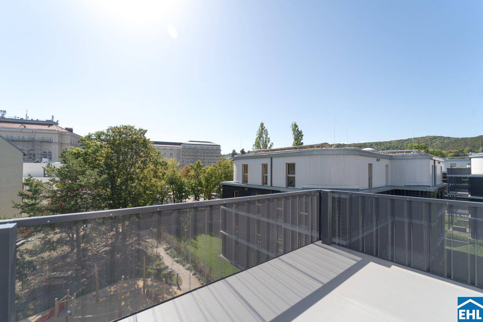 Heller Balkon mit Weitblick über moderne Architektur und grüne Bäume unter blauem Himmel.