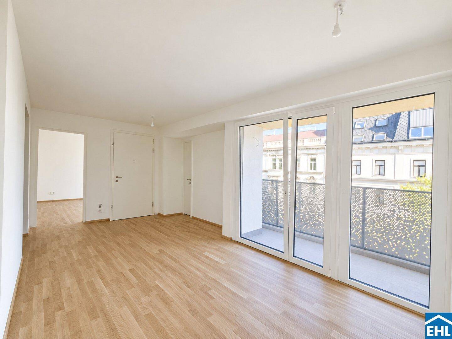 Geräumiger, heller Raum mit Holzboden und großen Fenstern, die zu einem Balkon führen.