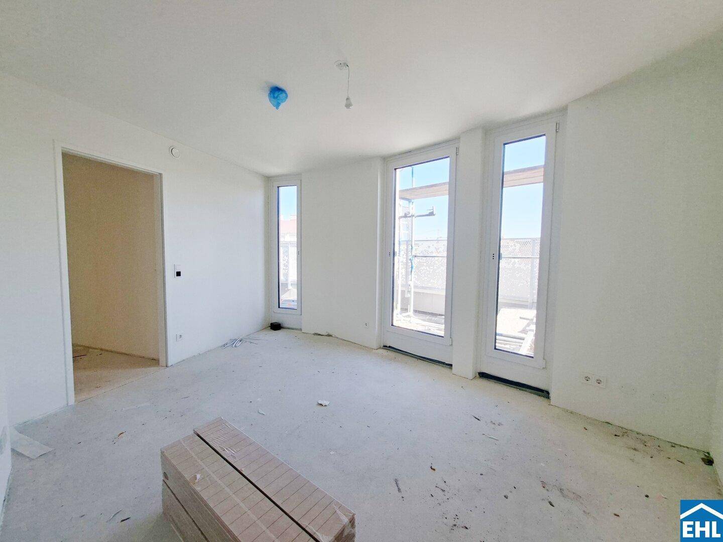 Heller Wohnraum im Rohbau mit großen Fenstern und offener Tür zu einem angrenzenden Zimmer.