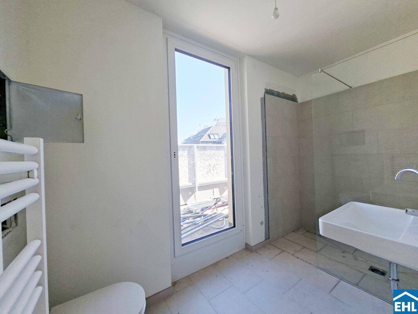 Unfertiges Badezimmer mit Fliesenboden, Waschbecken, Handtuchheizkörper und großem Fenster.