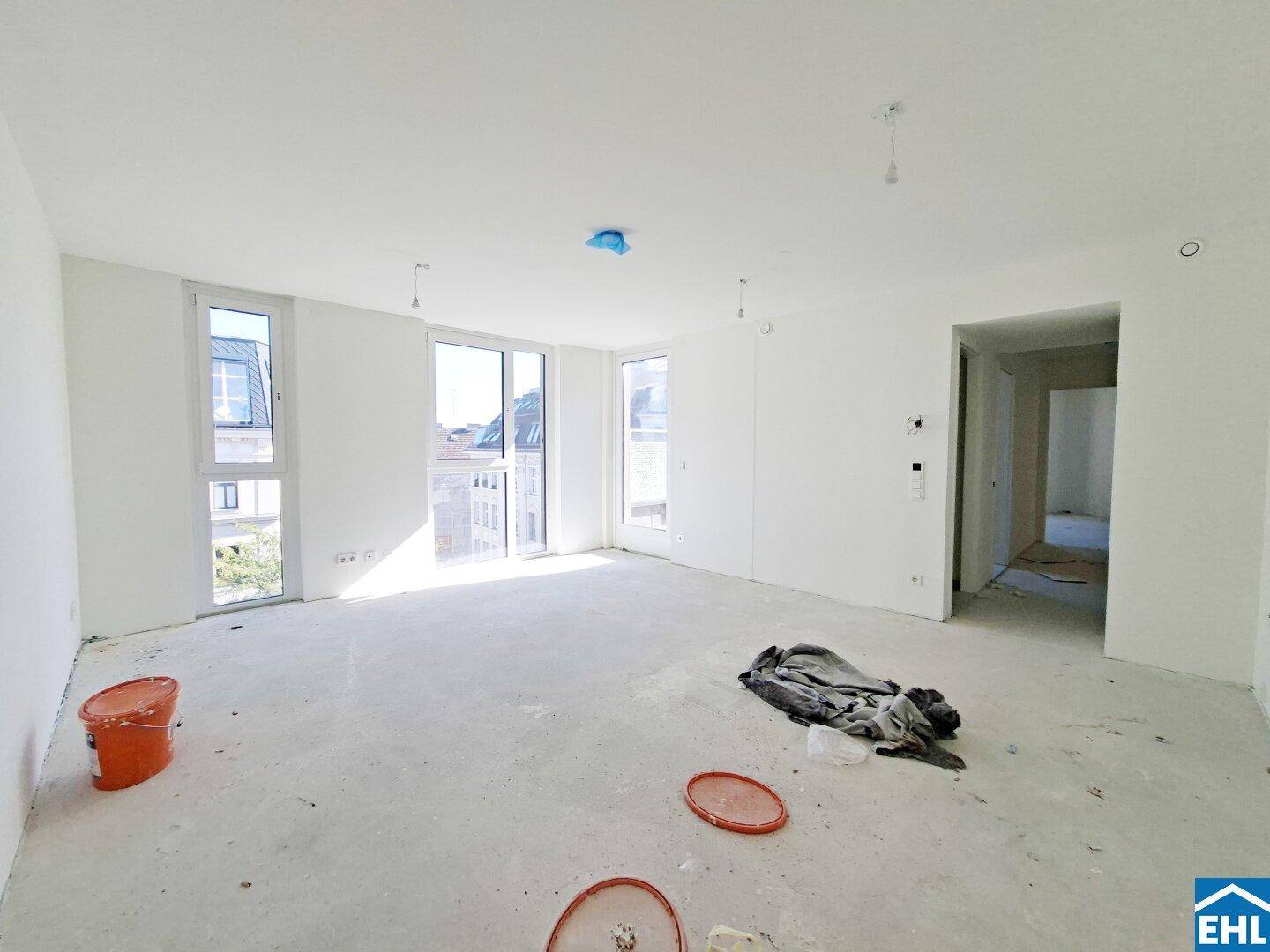 Geräumiger Wohnbereich im Rohbau mit viel Tageslicht, großen Fenstern und Zugang zu einem weiteren Zimmer.