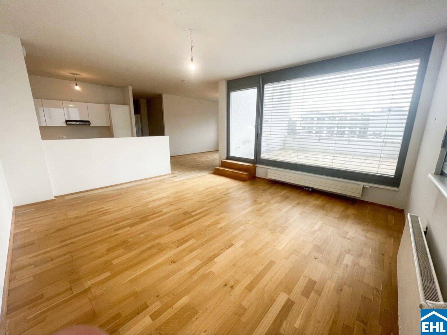 Heller Wohnbereich mit Holzboden, großen Fenstern und offener Küchenzeile im Hintergrund.