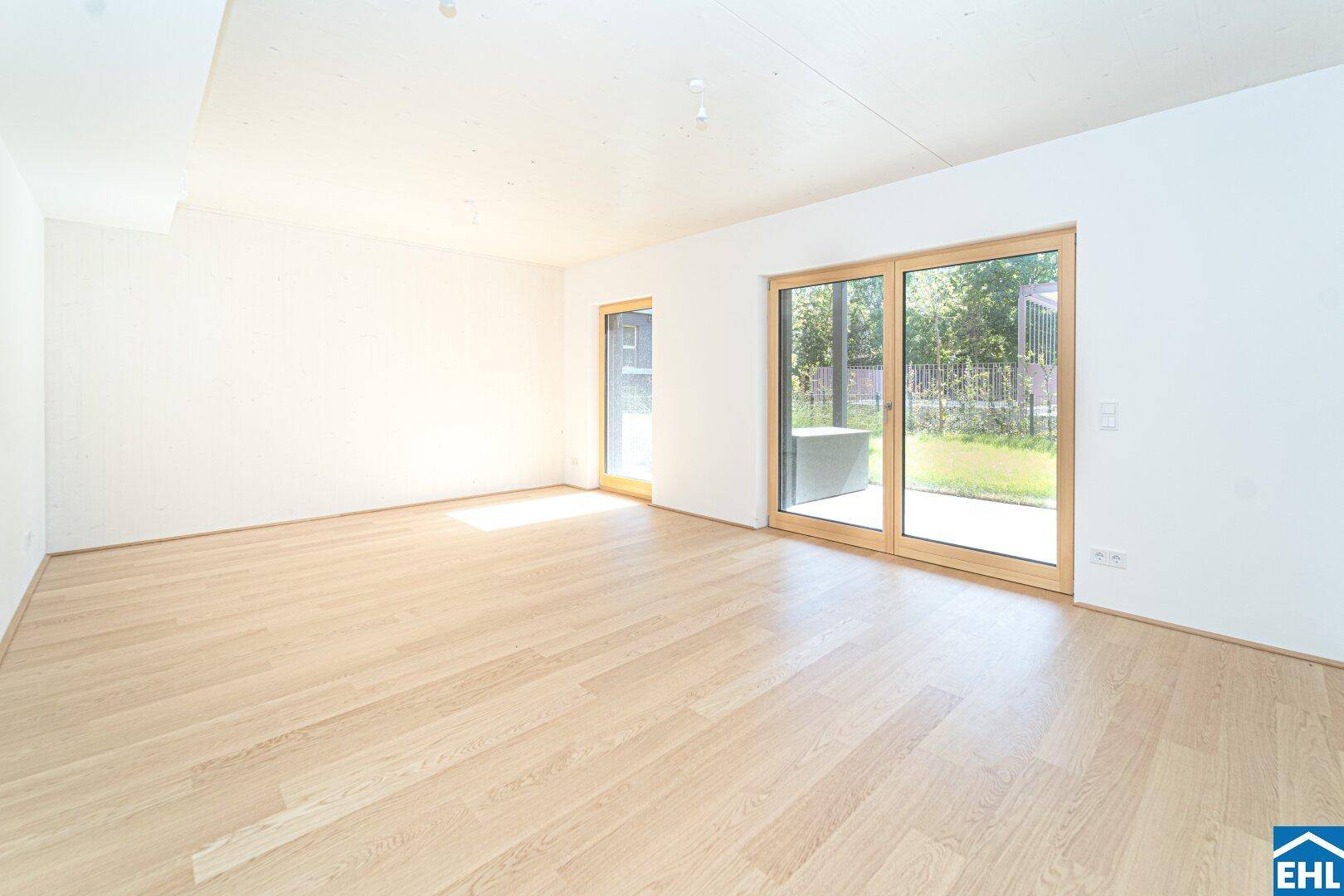 Helles, leeres Wohnzimmer mit großen Fenstern, Holzboden und Zugang zum Garten.