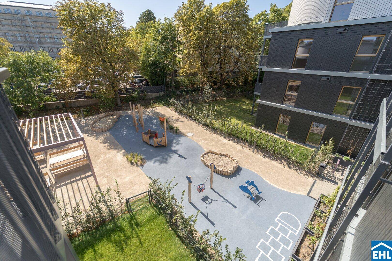 Vogelperspektive auf den modernen Spielplatz mit verschiedenen Spielgeräten und Sitzgelegenheiten zwischen den Gebäuden.