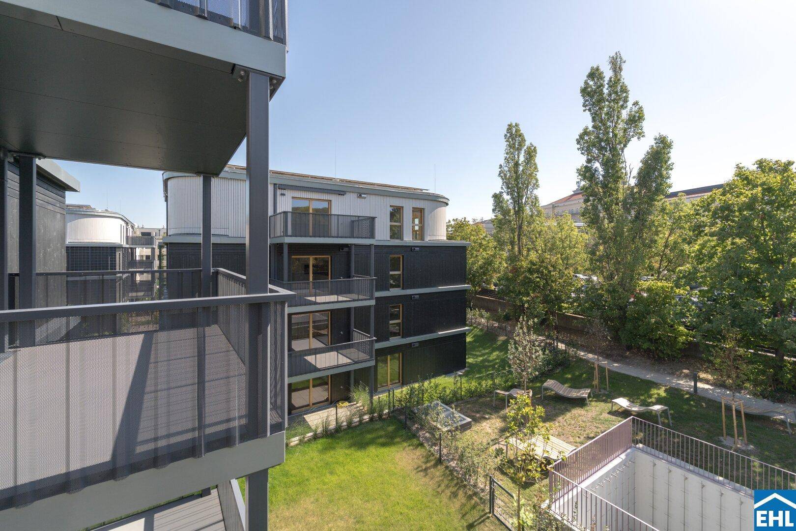 Blick auf die modernen Wohngebäude mit Balkonen und den gepflegten Gartenbereich mit Liegestühlen und Bäumen.