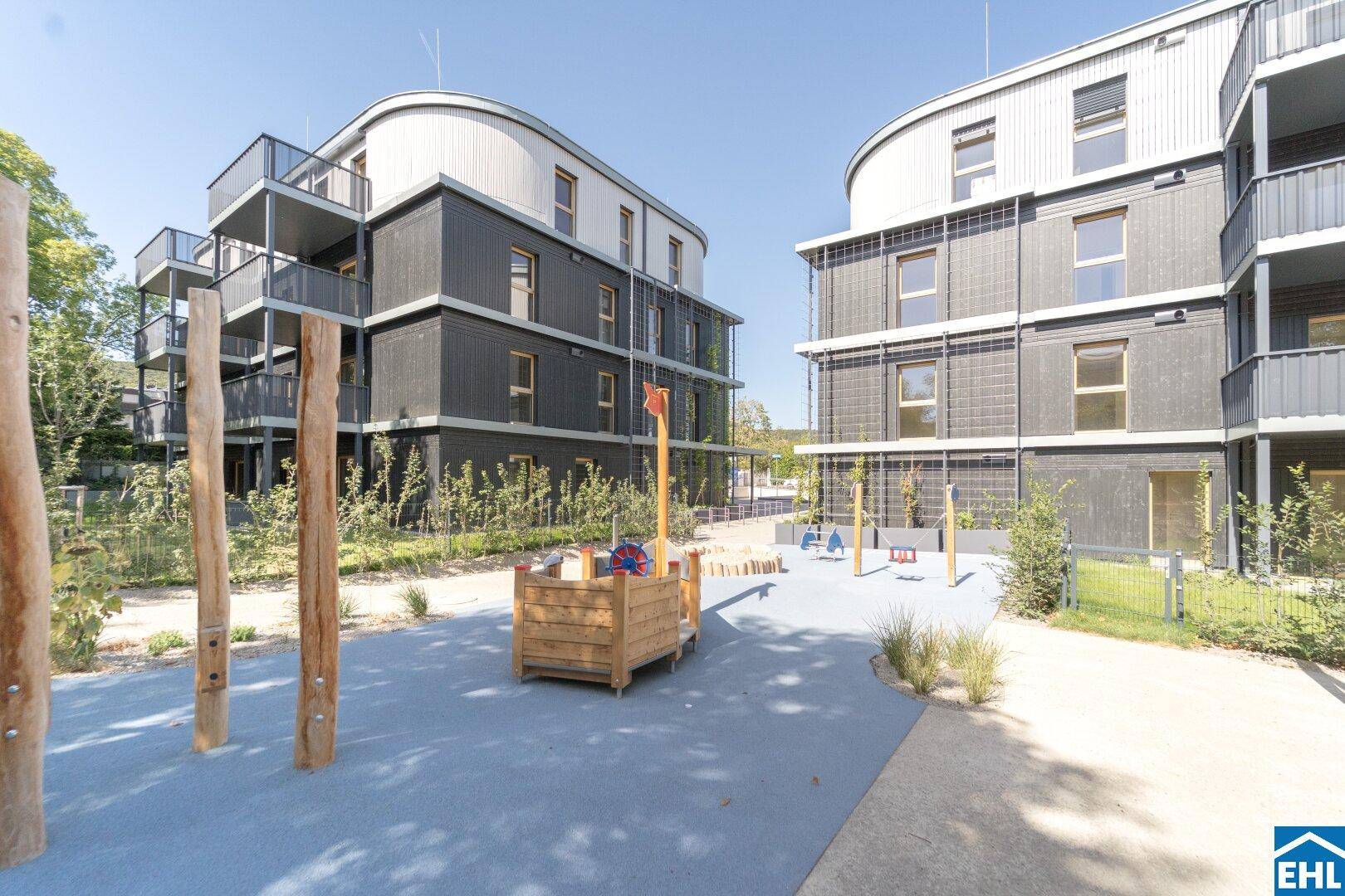 Weitläufiger Spielplatzbereich mit Holzelementen und zwei modernen Wohngebäuden im Hintergrund.