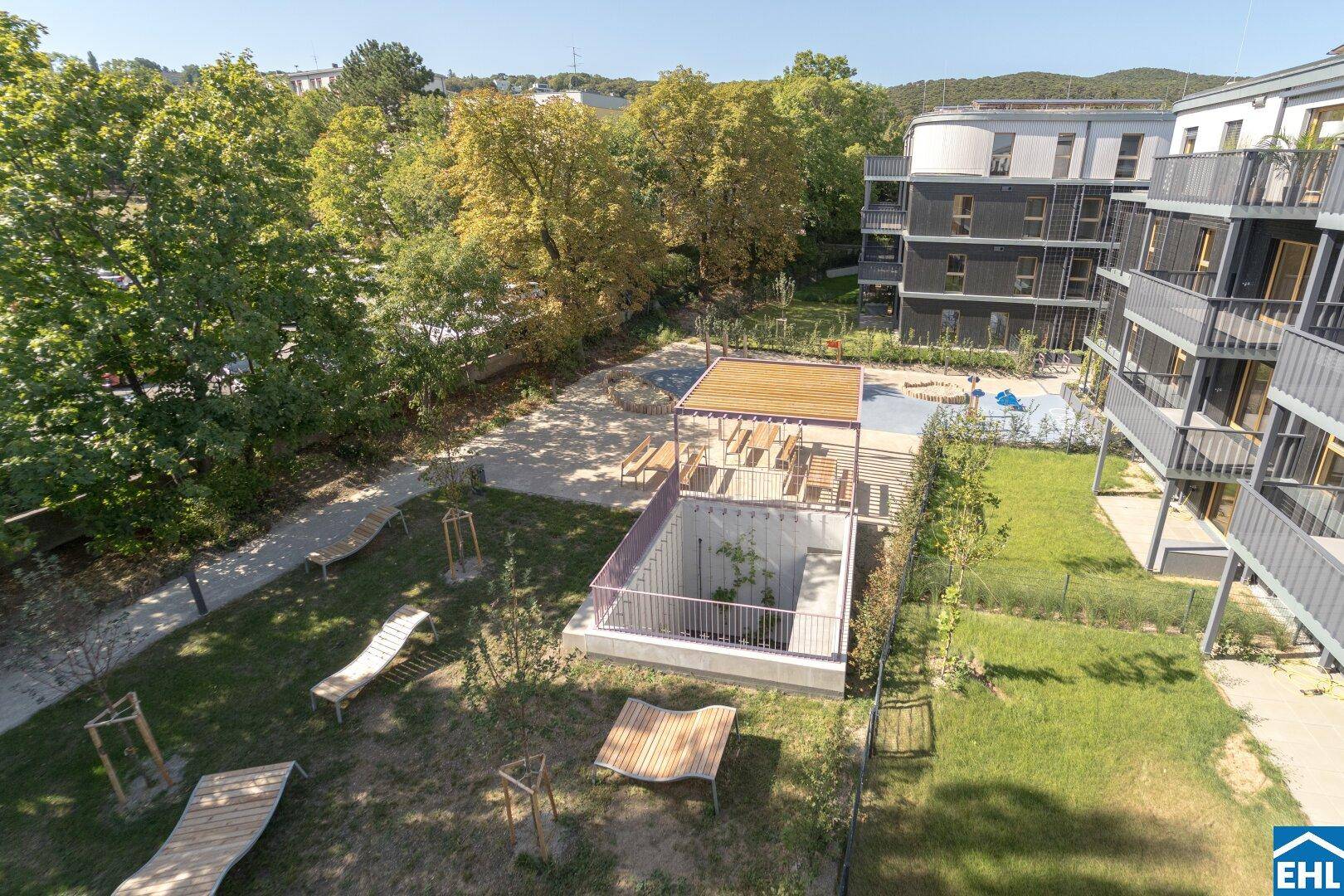 Weitläufiger Gartenbereich mit Spielplatz, Liegebänken und modernen Wohngebäuden unter strahlend blauem Himmel.