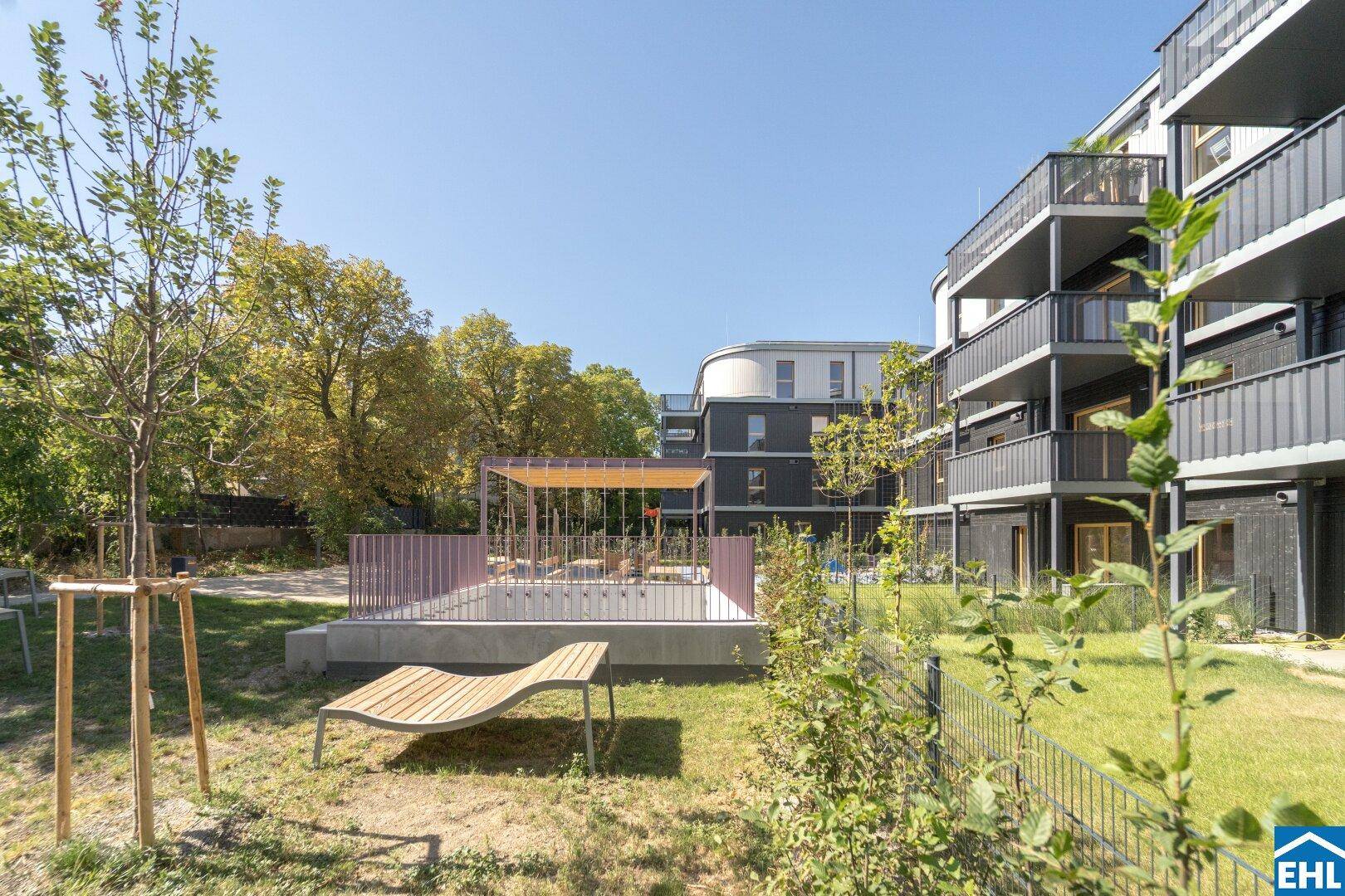 Ruhiger Gartenbereich mit Liegestuhl und junger Bepflanzung vor den modernen Gebäuden.