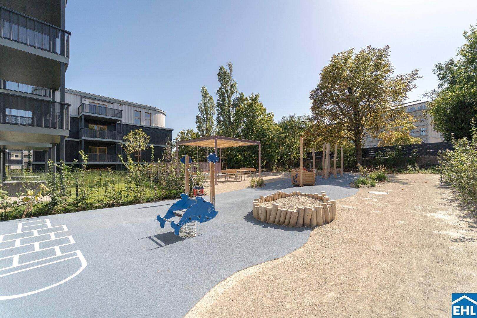 Sonniger Spielplatz mit Sandkasten und bunten Spielgeräten zwischen modernen Gebäuden.