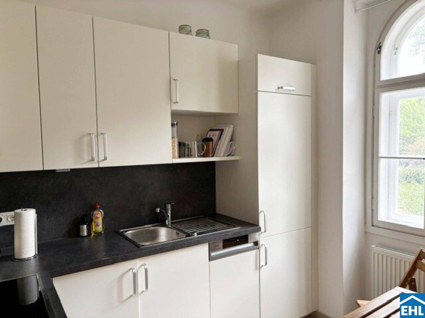 Moderne Einbauküche mit weißen Schränken, dunkler Arbeitsplatte, Spülbecken und Fenster.