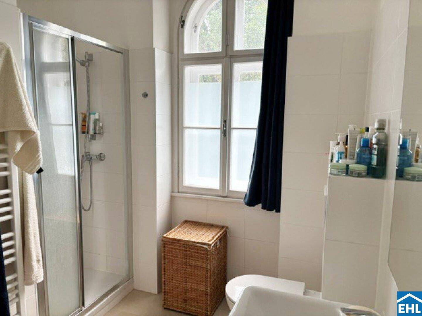 Badezimmer mit Duschkabine, Toilette, Fenster und beheiztem Handtuchhalter, hell gefliest.
