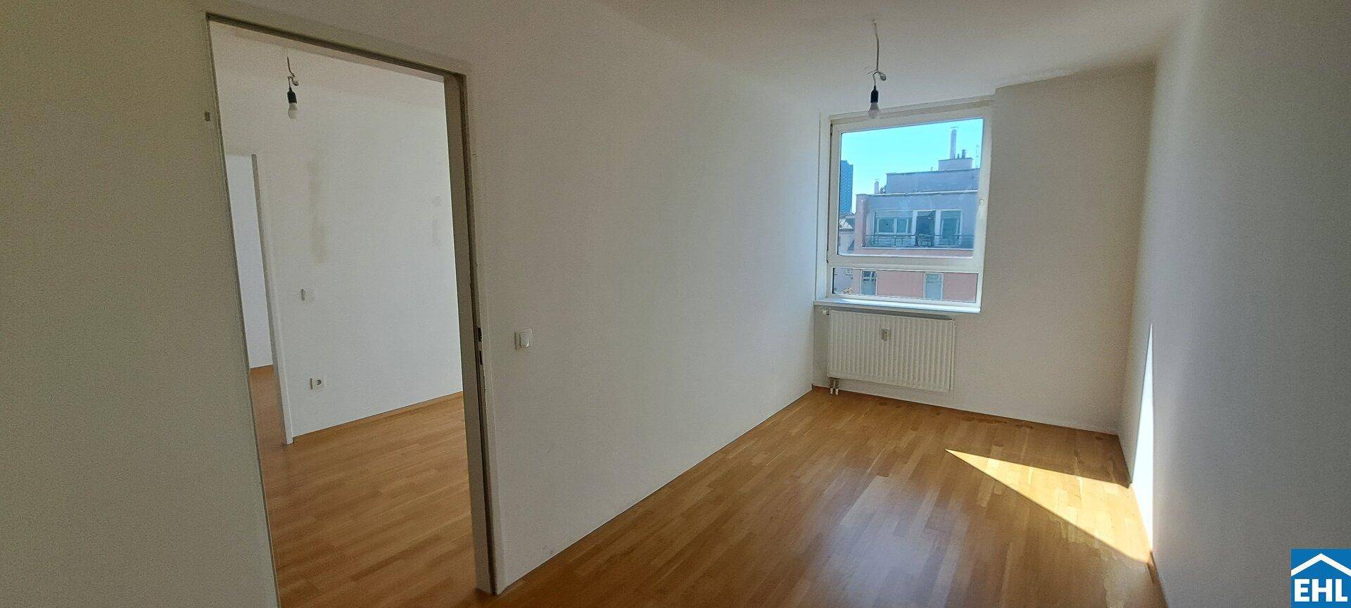 Leeres Zimmer mit Holzfußboden, großem Fenster und Ausblick auf die umliegenden Gebäude.