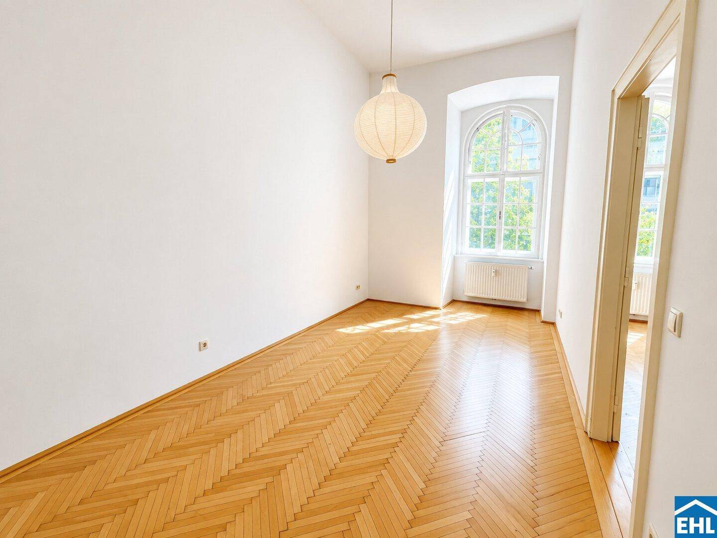 Heller Raum mit Fischgrätparkettboden, hohen Decken und großem Bogenfenster, das viel Licht hereinlässt.