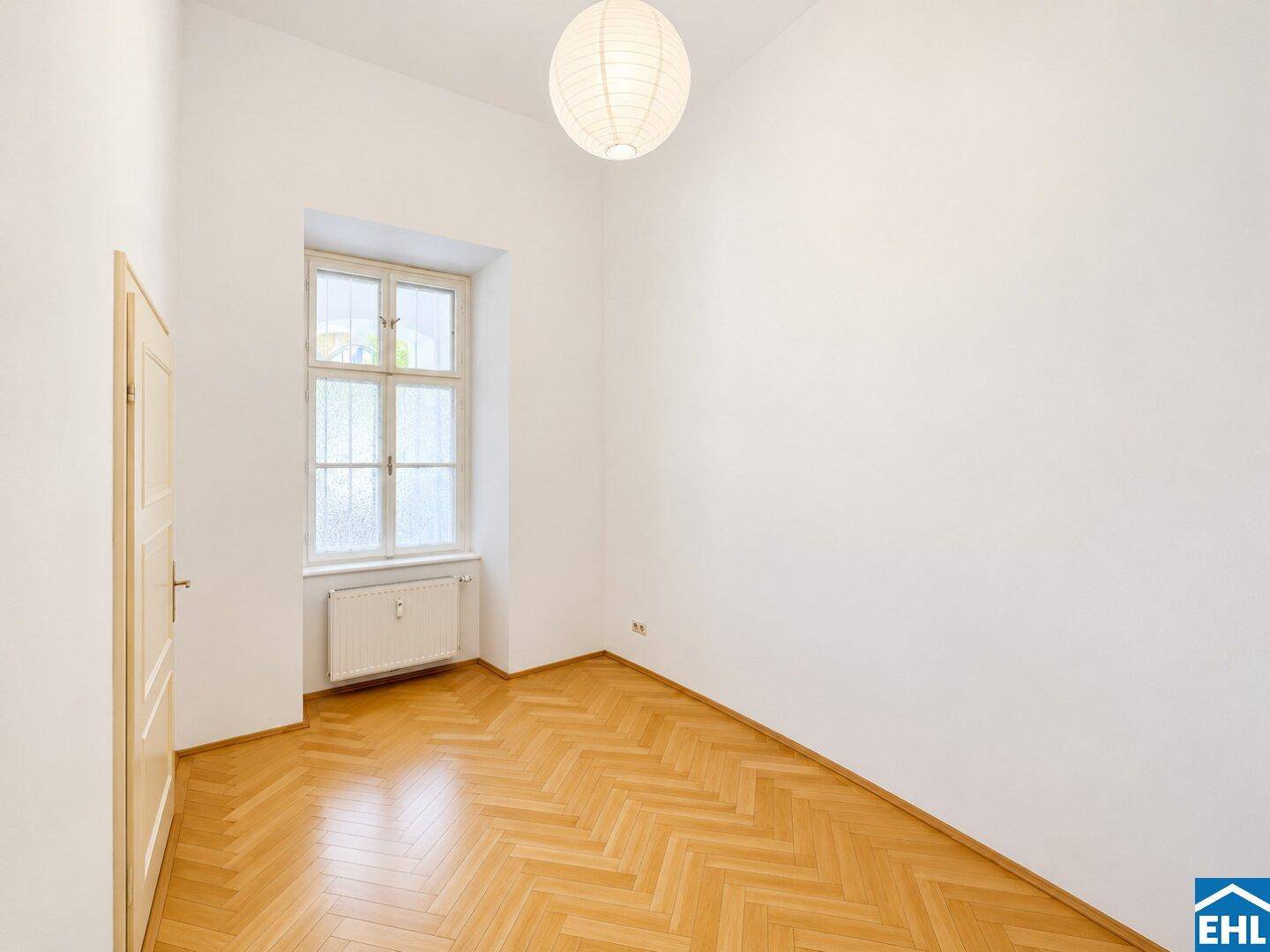 Gemütlicher Raum mit klassischem Fischgrätparkett, hohen Decken und einem Fenster mit Blick ins Grüne.