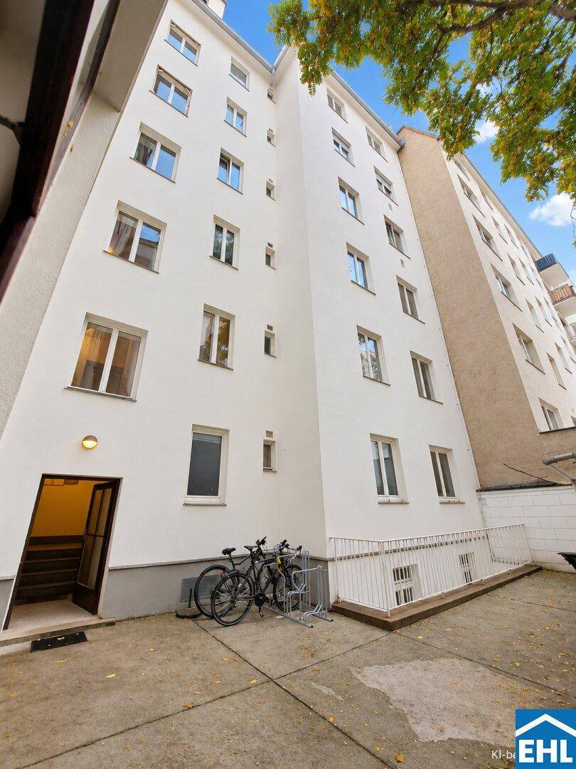 Weiße Gebäudefassade mit vielen Fenstern und Eingangsbereich, umgeben von Bäumen.