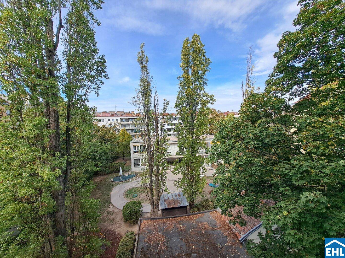 Grüner Ausblick auf einen Innenhof mit hohen Bäumen und einem Springbrunnen, umgeben von Gebäuden.