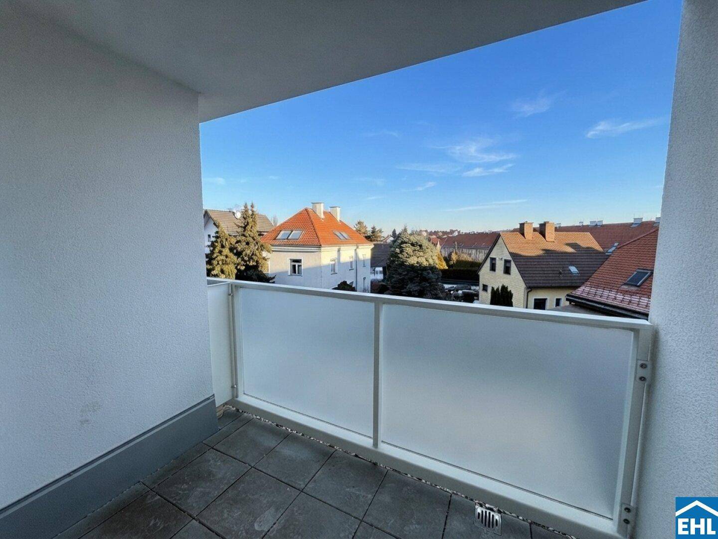 Heller Balkon mit Milchglasgeländer und Blick auf die umliegenden Häuser und den blauen Himmel.