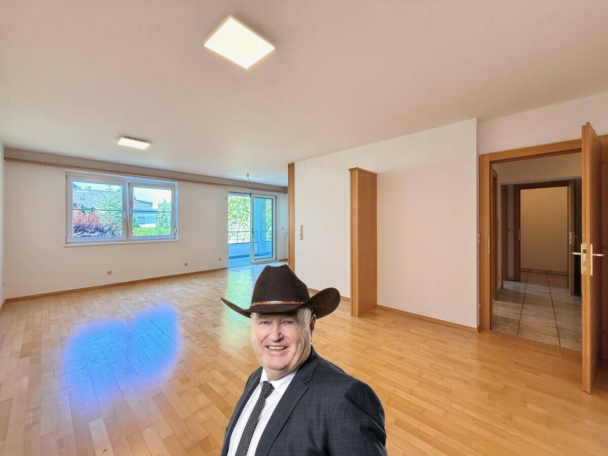 Helles, leeres Wohnzimmer mit Parkettboden und Zugang zu einem Balkon.