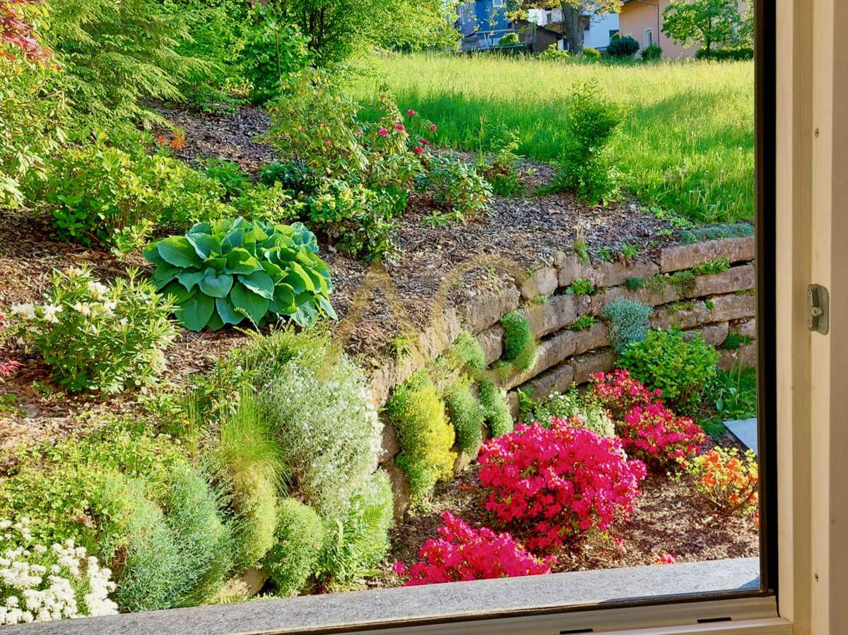 Blick aus dem Fenster auf einen farbenfrohen Garten mit Blumenbeeten und Steinmauer.