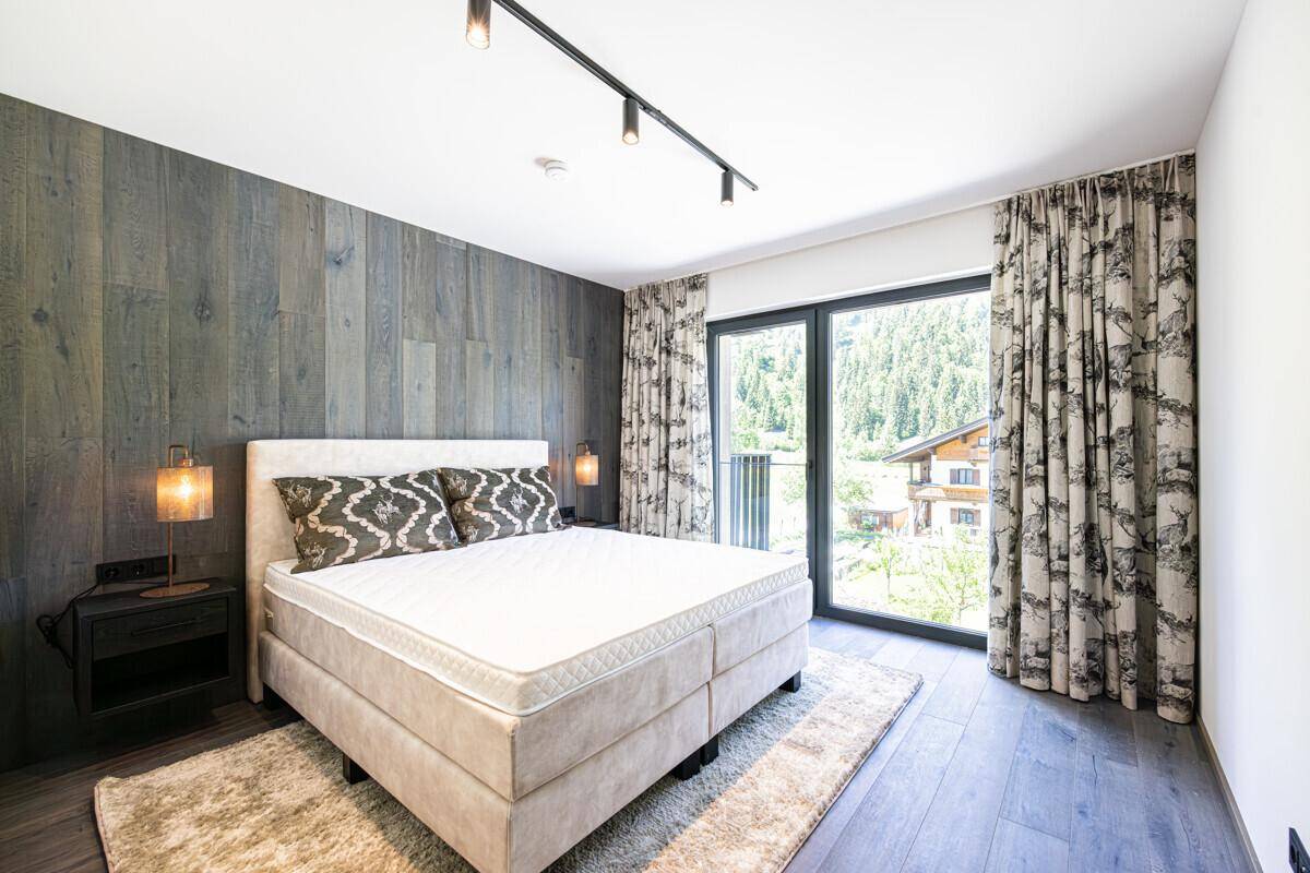 Helles Schlafzimmer mit modernem Bett, Holzwand und großem Fenster mit Blick ins Grüne.
