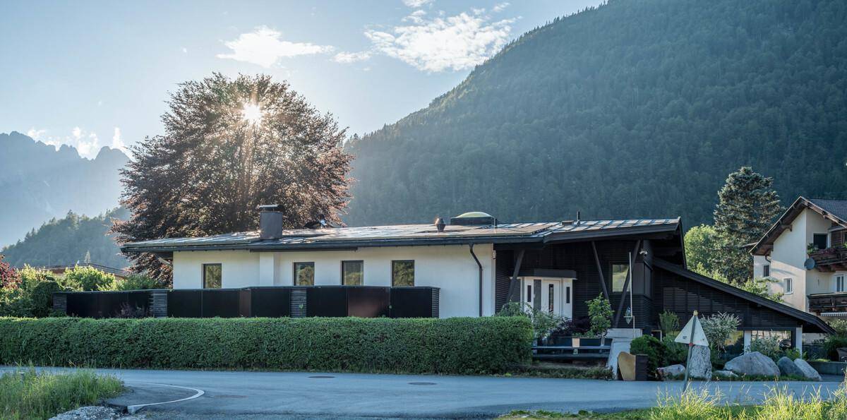 Modernes Haus mit dunkler Holzfassade und gepflegter Hecke vor alpiner Bergkulisse.