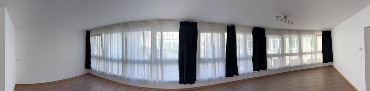 Panoramaansicht eines hellen Wohnzimmers mit vielen Fenstern und Vorhängen.