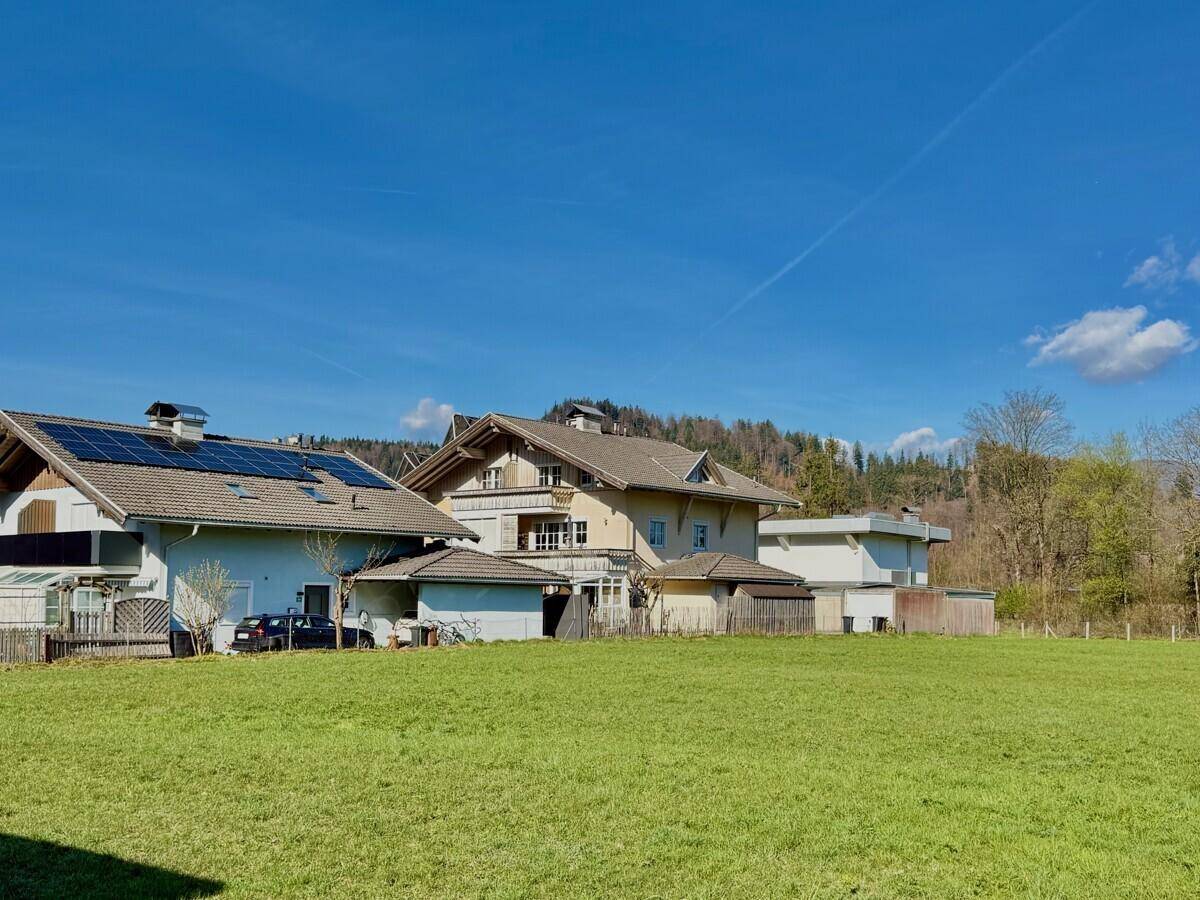 Weitläufige grüne Landschaft mit Wohnhäusern und bewaldeten Hügeln im Hintergrund.