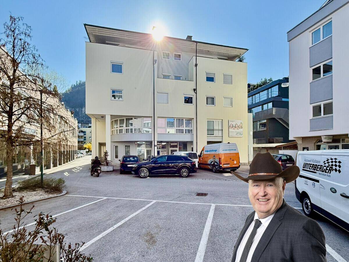 Modernes Gebäude mit Parkplätzen davor, beleuchtet von der Sonne, mit einer Person im Vordergrund.