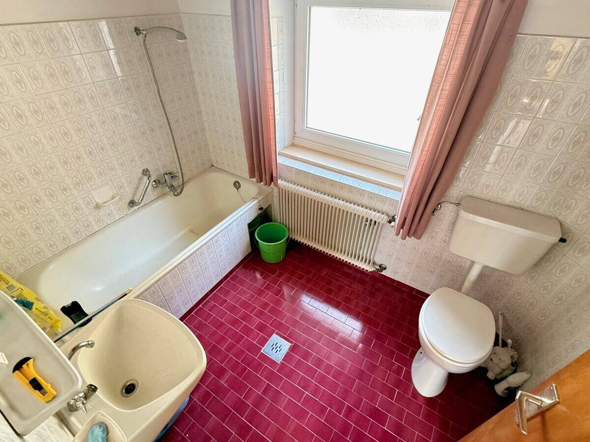Badezimmer im 80er-Jahre-Stil mit Badewanne, WC, Waschbecken und roten Bodenfliesen.