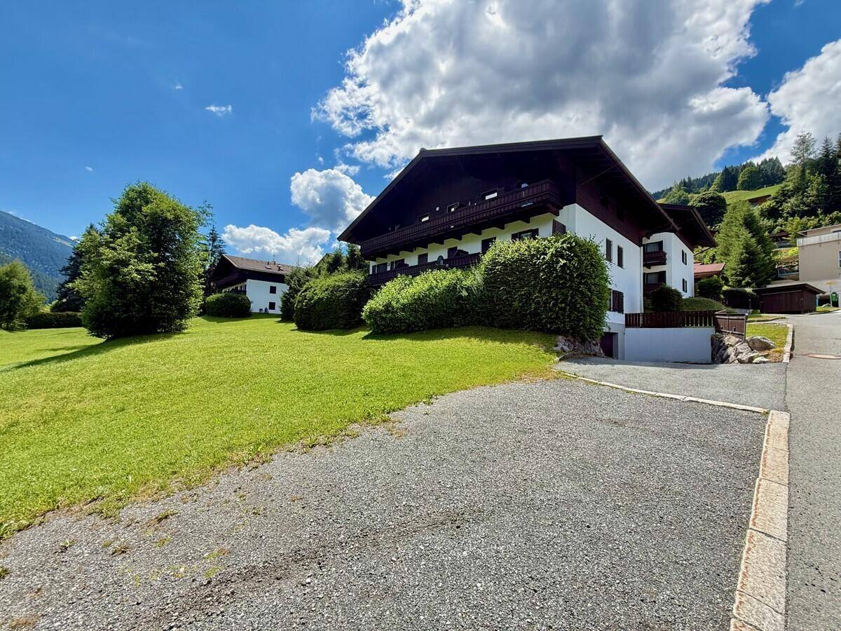 Mehrfamilienhaus im alpinen Stil mit großem Rasen und Schotterparkplatz.