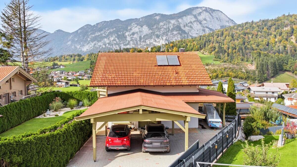 Einfamilienhaus mit Carport, Ziegeldach und Photovoltaikanlage vor einer beeindruckenden Bergkulisse.