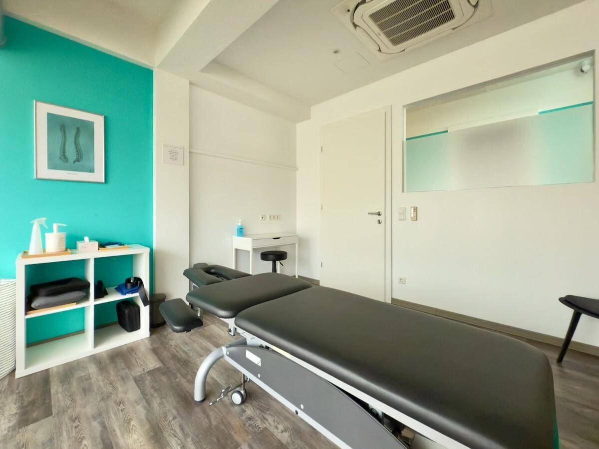 Heller Behandlungsraum mit Therapieliege, türkiser Akzentwand und Fenster zur Rezeption.