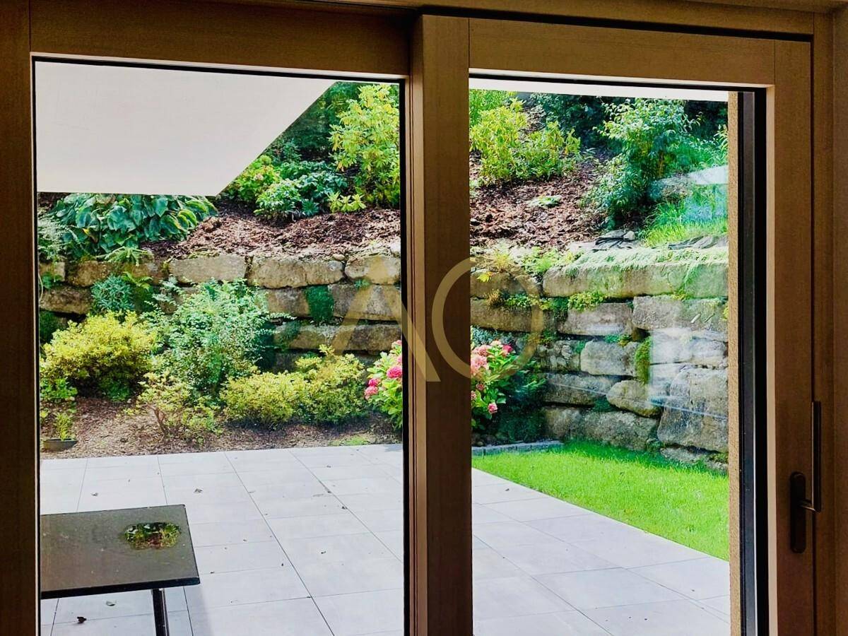 Blick von innen auf eine gepflasterte Terrasse und den grünen, terrassierten Garten.