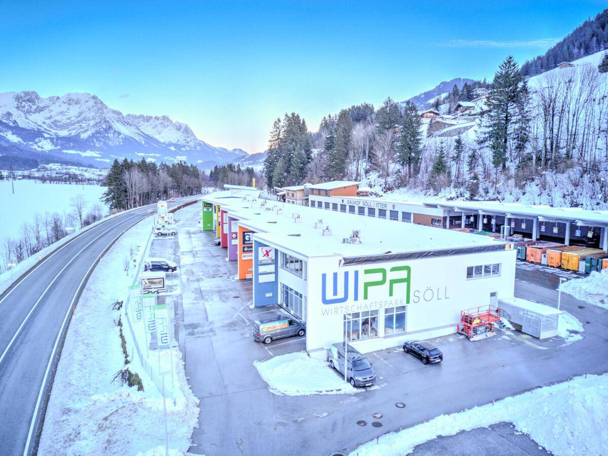 Luftaufnahme eines modernen Gewerbeparks in verschneiter Winterlandschaft mit beeindruckendem Bergpanorama.