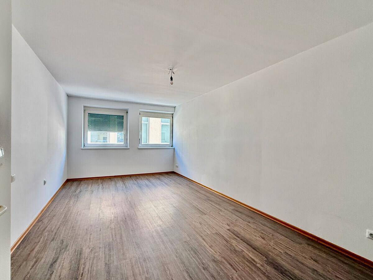 Leeres Zimmer mit Laminatboden, weißen Wänden und zwei Fenstern mit Rollläden.